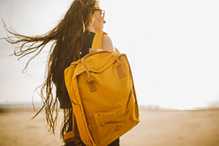 Werbe-Rucksäcke bedrucken Sie bei laprinta Eine Frau mit langen braunen Haaren steht mit dem Rücken zum Betrachter und trägt einen senfgelben Rucksack