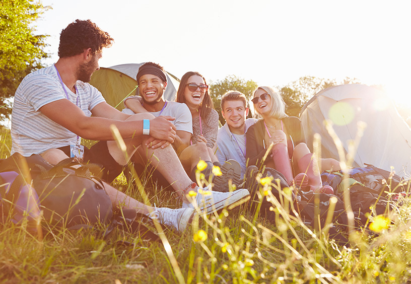 Fesival Werbeartikel bedrucken bei laprinta Eine Gruppe aus Männern und Frauen sitzen in der Wiese und lachen, im Hintergrund ist ein kleines Zelt